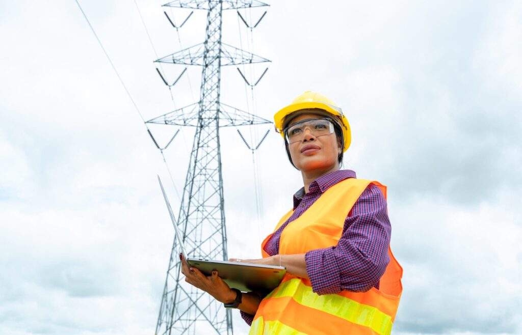 A female engineer stands in front of an electrical tower with her laptop.