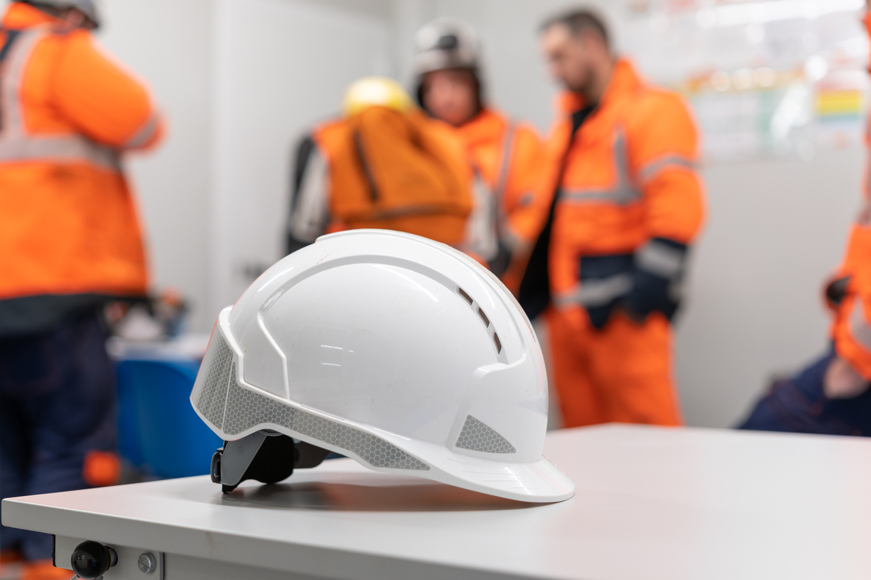 White safety helmet on a table with construction crew in high-visibility gear blurred in the background, symbolizing workplace safety protocols and change management.