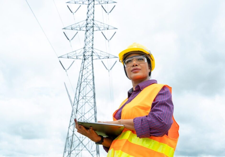 A female engineer stands in front of an electrical tower with her laptop.