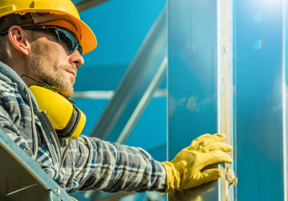Caucasian Construction Industry Worker Wearing Yellow Hard Hat and Sunglasses Staying Next to Aluminium Building Frame.